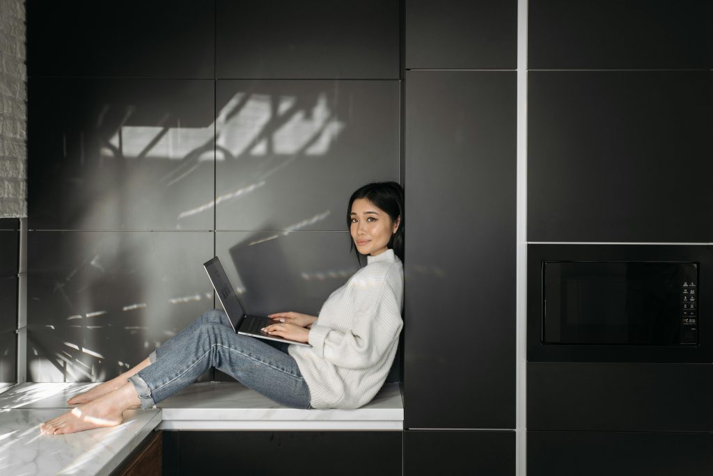 Young woman sitting on countertop using laptop for remote work in stylish kitchen.